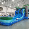 Large tropical-themed inflatable combo featuring dual blue water slides flanked by inflatable palm trees. The structure includes a tall climbing wall with blue and green panels, leading to the slide entrance. A spacious splash pool area is visible at the base. Set up in a large indoor warehouse with white walls and metal roofing, showcasing its size and commercial use.