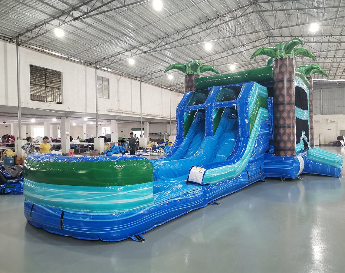 Large tropical-themed inflatable combo featuring dual blue water slides flanked by inflatable palm trees. The structure includes a tall climbing wall with blue and green panels, leading to the slide entrance. A spacious splash pool area is visible at the base. Set up in a large indoor warehouse with white walls and metal roofing, showcasing its size and commercial use.