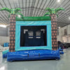 Rear view of Island Drop Dual Lane Commercial Inflatable Combo (Wet/Dry) in a warehouse setting. The inflatable features two large brown palm tree pillars with green leaves, a teal-colored entrance frame, and a blue base. The interior of the bounce house is visible, showing inflatable obstacles. A person can be seen in the background, providing scale to the large structure.