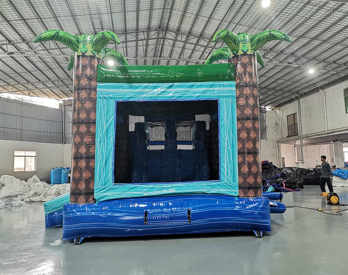 Rear view of Island Drop Dual Lane Commercial Inflatable Combo (Wet/Dry) in a warehouse setting. The inflatable features two large brown palm tree pillars with green leaves, a teal-colored entrance frame, and a blue base. The interior of the bounce house is visible, showing inflatable obstacles. A person can be seen in the background, providing scale to the large structure.