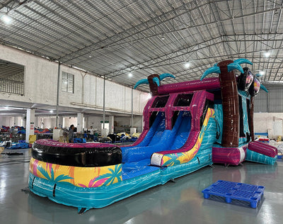 Three-quarter view of Island Flow Dual Lane Commercial Inflatable Combo in a spacious warehouse. The inflatable displays a tropical theme with large sunglasses, dual blue slides, palm tree pillars, and a colorful splash pool area. The warehouse interior is visible with metal roofing, support beams, and other inflatable products, demonstrating its size and commercial application.