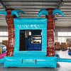 Large inflatable tropical-themed combo featuring a bounce house with palm tree decorations, attached slide, and splash pool. The structure has vibrant turquoise, pink, and yellow colors with brown palm tree trunks and a large inflatable sunglasses decoration on top. Set up in an indoor warehouse with metal roofing and other inflatable products visible in the background.