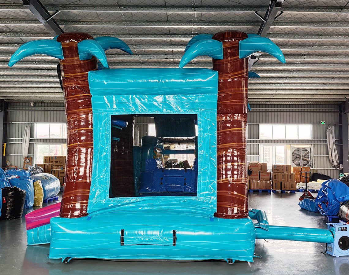 Large inflatable tropical-themed combo featuring a bounce house with palm tree decorations, attached slide, and splash pool. The structure has vibrant turquoise, pink, and yellow colors with brown palm tree trunks and a large inflatable sunglasses decoration on top. Set up in an indoor warehouse with metal roofing and other inflatable products visible in the background.