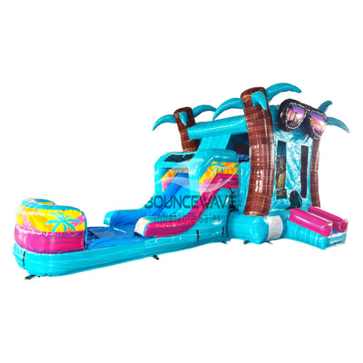 Large inflatable tropical-themed combo featuring a bounce house with palm tree decorations, attached slide, and splash pool. The structure has vibrant turquoise, pink, and yellow colors with brown palm tree trunks. Set up against a white background.