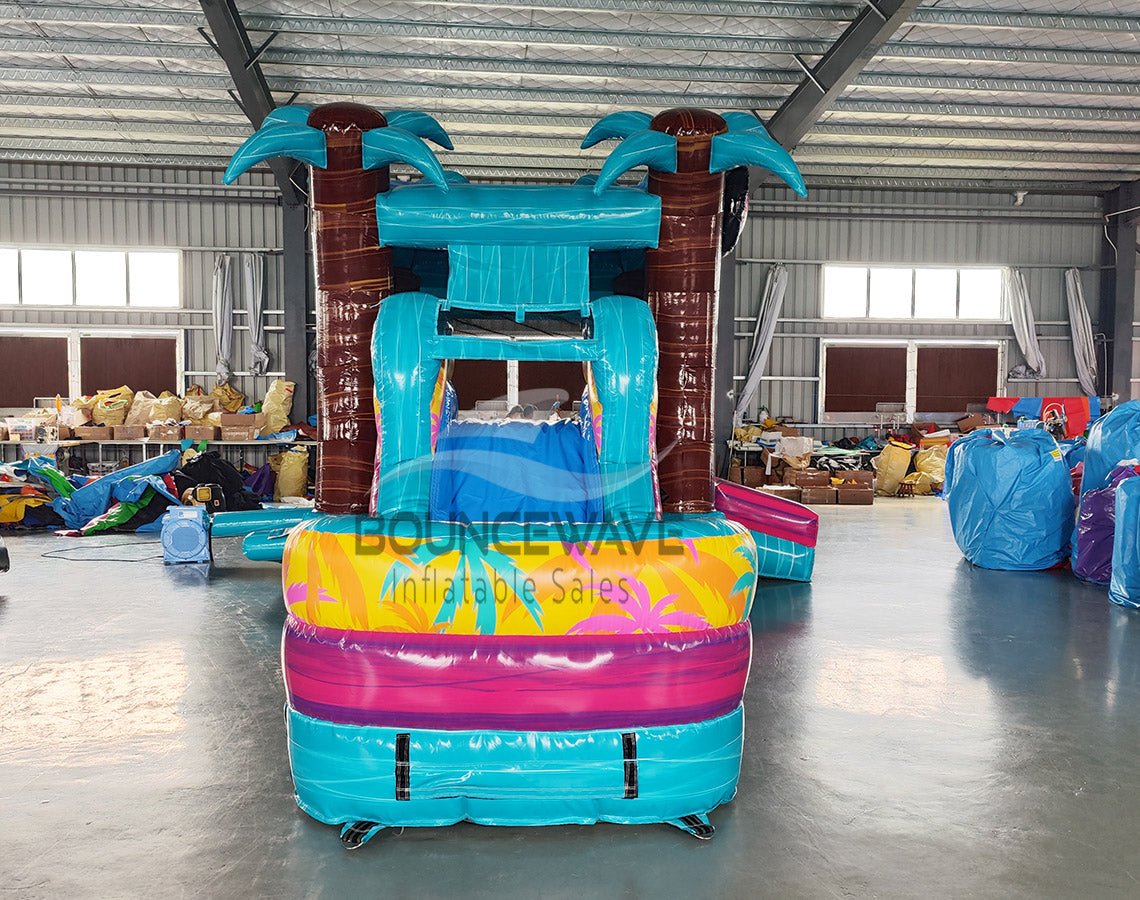 Front view of Island Flow Econo Combo featuring turquoise bounce house with palm trees, colorful slide, and splash pool. Set up in an indoor facility with various other inflatable products and equipment visible in the background.