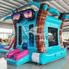 Front view of Island Flow Econo Combo, showcasing the bounce house entrance with palm tree decorations, colorful slide, and splash pool area. Set up in an indoor warehouse with metal roofing and other inflatable products visible in the background.