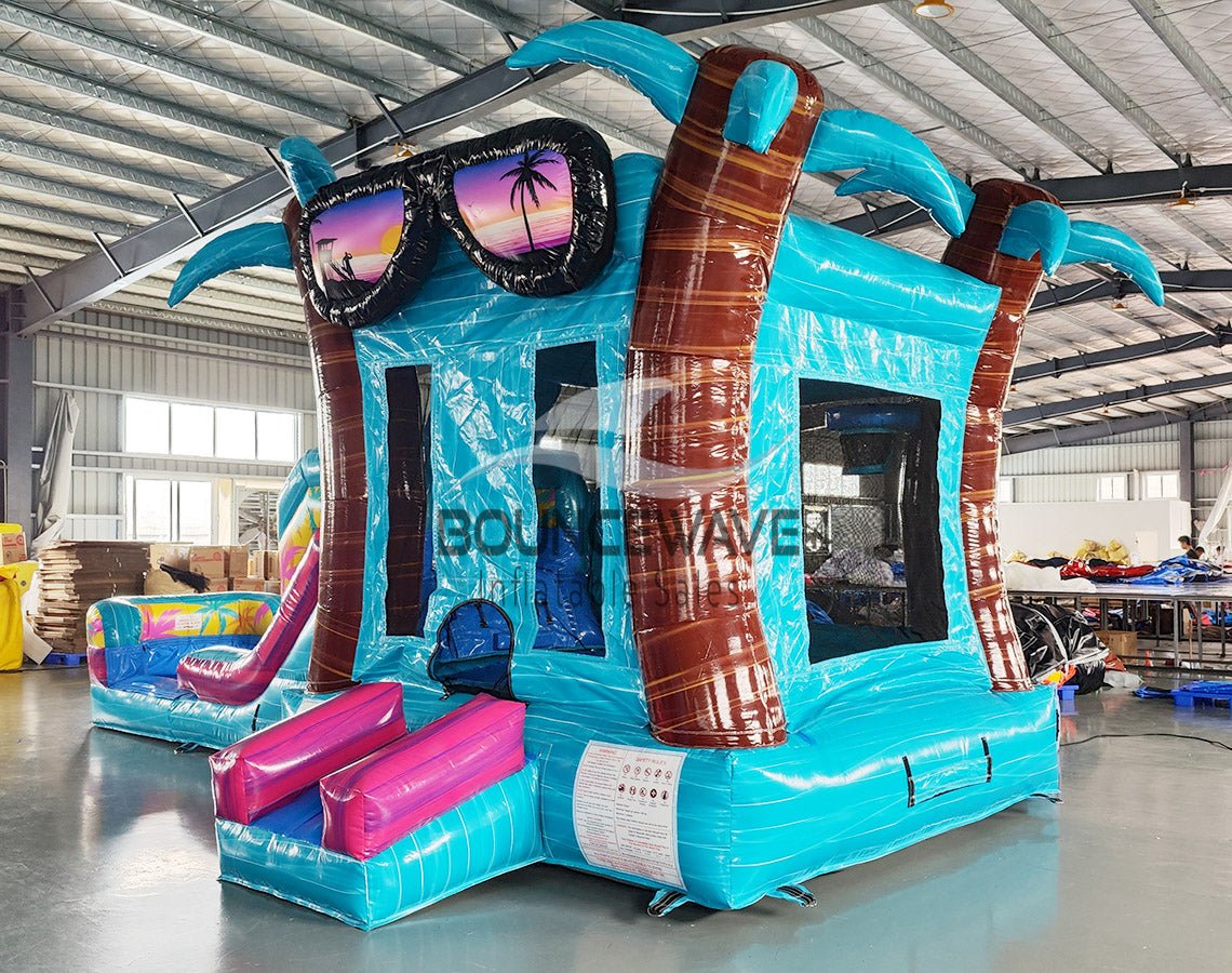 Front view of Island Flow Econo Combo, showcasing the bounce house entrance with palm tree decorations, colorful slide, and splash pool area. Set up in an indoor warehouse with metal roofing and other inflatable products visible in the background.