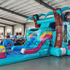 Wide-angle view of Island Flow Econo Combo showing bright turquoise, pink, and yellow design with inflatable palm trees, curved slide, and large splash pool area. The combo is set up in a spacious indoor warehouse with metal roofing, bright lighting, and other equipment visible.