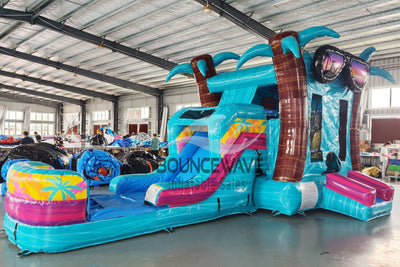 Wide-angle view of Island Flow Econo Combo showing bright turquoise, pink, and yellow design with inflatable palm trees, curved slide, and large splash pool area. The combo is set up in a spacious indoor warehouse with metal roofing, bright lighting, and other equipment visible.