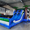 Three-quarter view of Island Thunder Dual Lane Commercial Inflatable Combo (Wet/Dry) in a warehouse setting. The inflatable displays dual blue water slides, palm tree pillars, a white bounce house area, and a large blue splash pool. Colorful inflatables and storage bags are visible in the background, showcasing its commercial context.