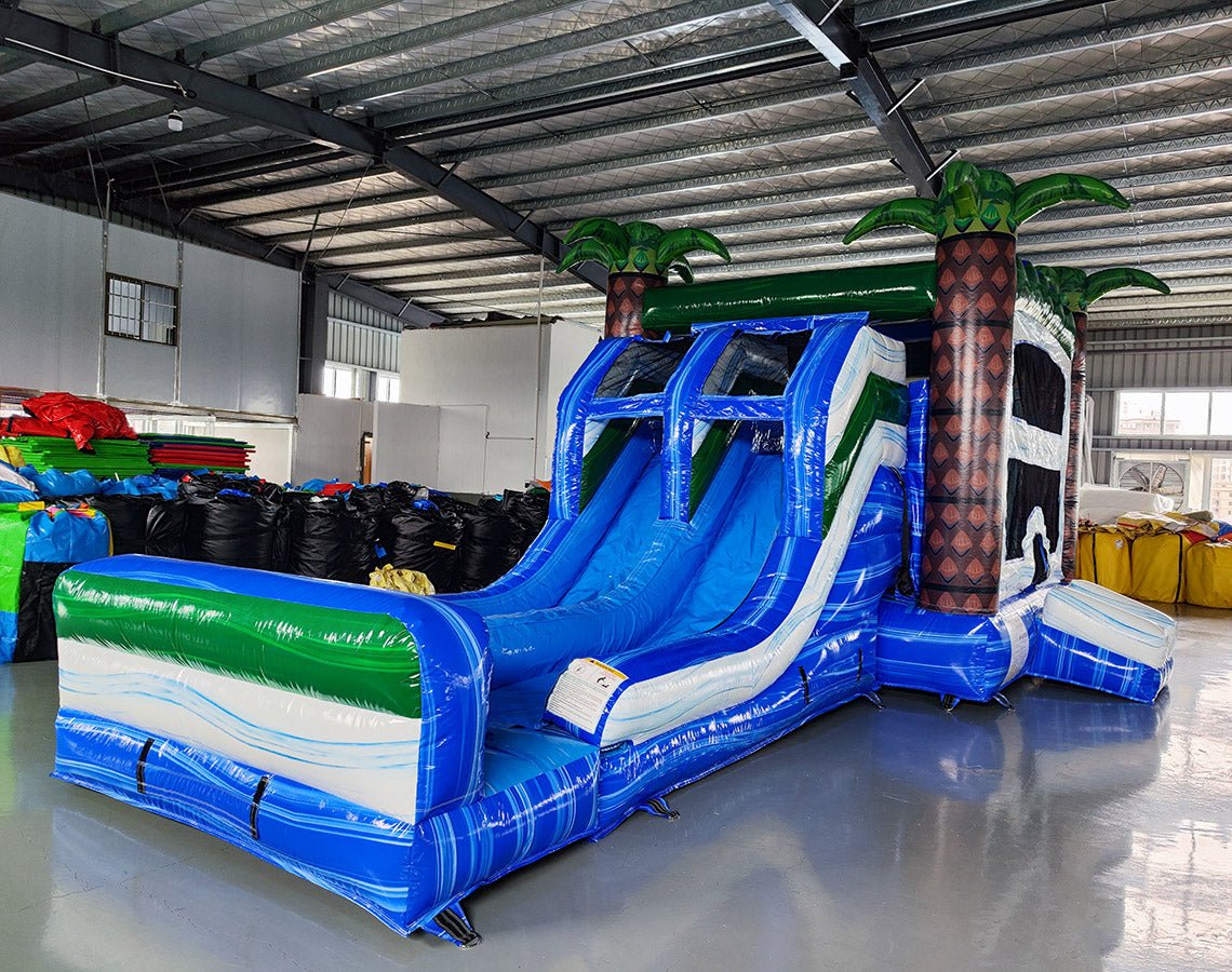 Three-quarter view of Island Thunder Dual Lane Commercial Inflatable Combo (Wet/Dry) in a warehouse setting. The inflatable displays dual blue water slides, palm tree pillars, a white bounce house area, and a large blue splash pool. Colorful inflatables and storage bags are visible in the background, showcasing its commercial context.