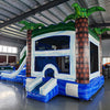 cross rear view of Island Thunder Dual Lane Commercial Inflatable Combo (Wet/Dry) featuring a tropical theme with inflatable palm trees. The structure has a white bounce house area with mesh windows, dual blue water slides, and a blue splash pool base. Set up in a large indoor warehouse with metal roofing and various equipment visible in the background.