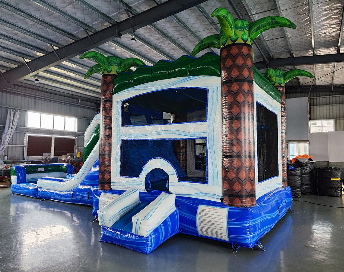 cross rear view of Island Thunder Dual Lane Commercial Inflatable Combo (Wet/Dry) featuring a tropical theme with inflatable palm trees. The structure has a white bounce house area with mesh windows, dual blue water slides, and a blue splash pool base. Set up in a large indoor warehouse with metal roofing and various equipment visible in the background.