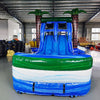 Front view of Island Thunder Dual Lane Commercial Inflatable Combo (Wet/Dry) featuring a tropical theme with inflatable palm trees. The structure has dual blue water slides, a white bounce house area with mesh windows, and a large blue splash pool base. Inflatable palm trees with brown trunks and green leaves flank the sides. Set up in a spacious indoor warehouse with metal roofing, fluorescent lighting, and various other inflatable products visible in the background.