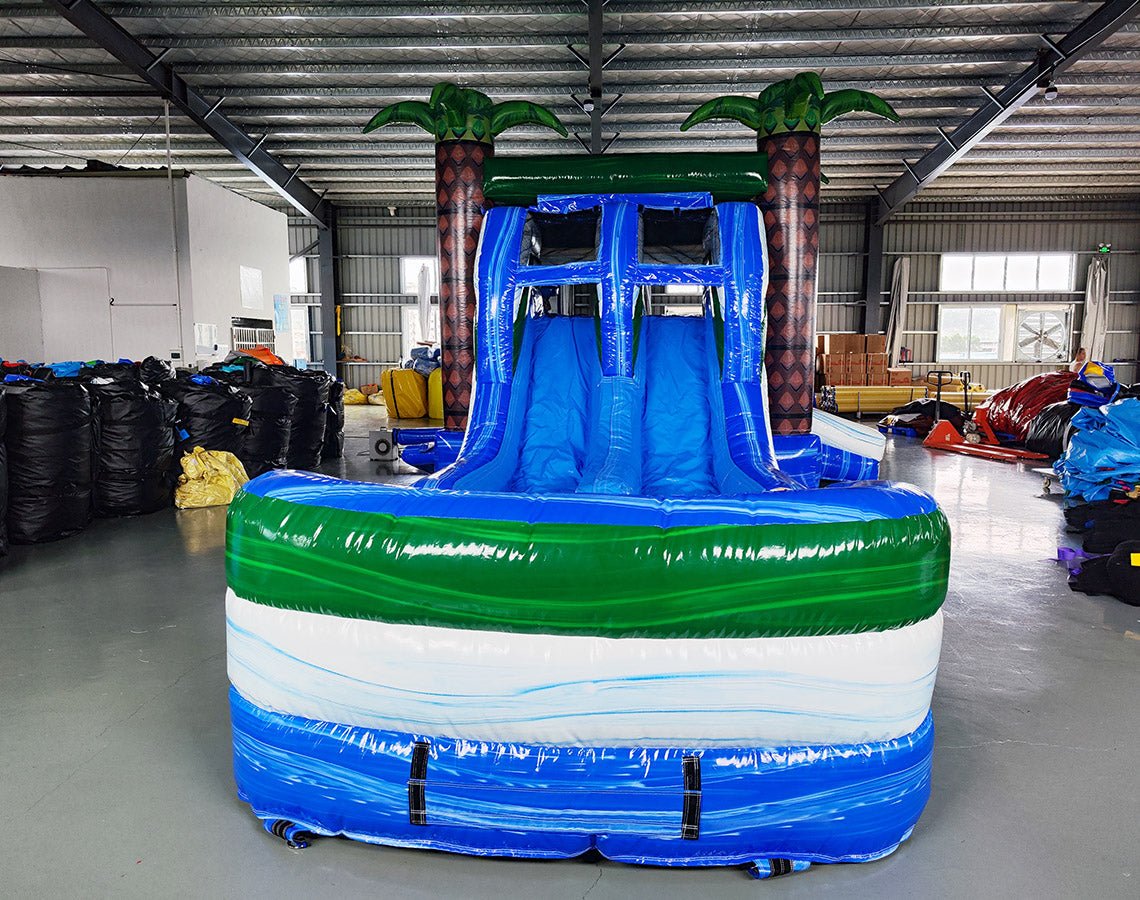 Front view of Island Thunder Dual Lane Commercial Inflatable Combo (Wet/Dry) featuring a tropical theme with inflatable palm trees. The structure has dual blue water slides, a white bounce house area with mesh windows, and a large blue splash pool base. Inflatable palm trees with brown trunks and green leaves flank the sides. Set up in a spacious indoor warehouse with metal roofing, fluorescent lighting, and various other inflatable products visible in the background.