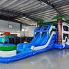 Side view of Island Thunder Dual Lane Commercial Inflatable Combo (Wet/Dry) showing dual blue water slides, inflatable palm trees with brown trunks and green leaves, and a large blue splash pool. The combo includes a white bounce house area and green accents. Set in a warehouse with metal ceiling and various inflatable products visible.