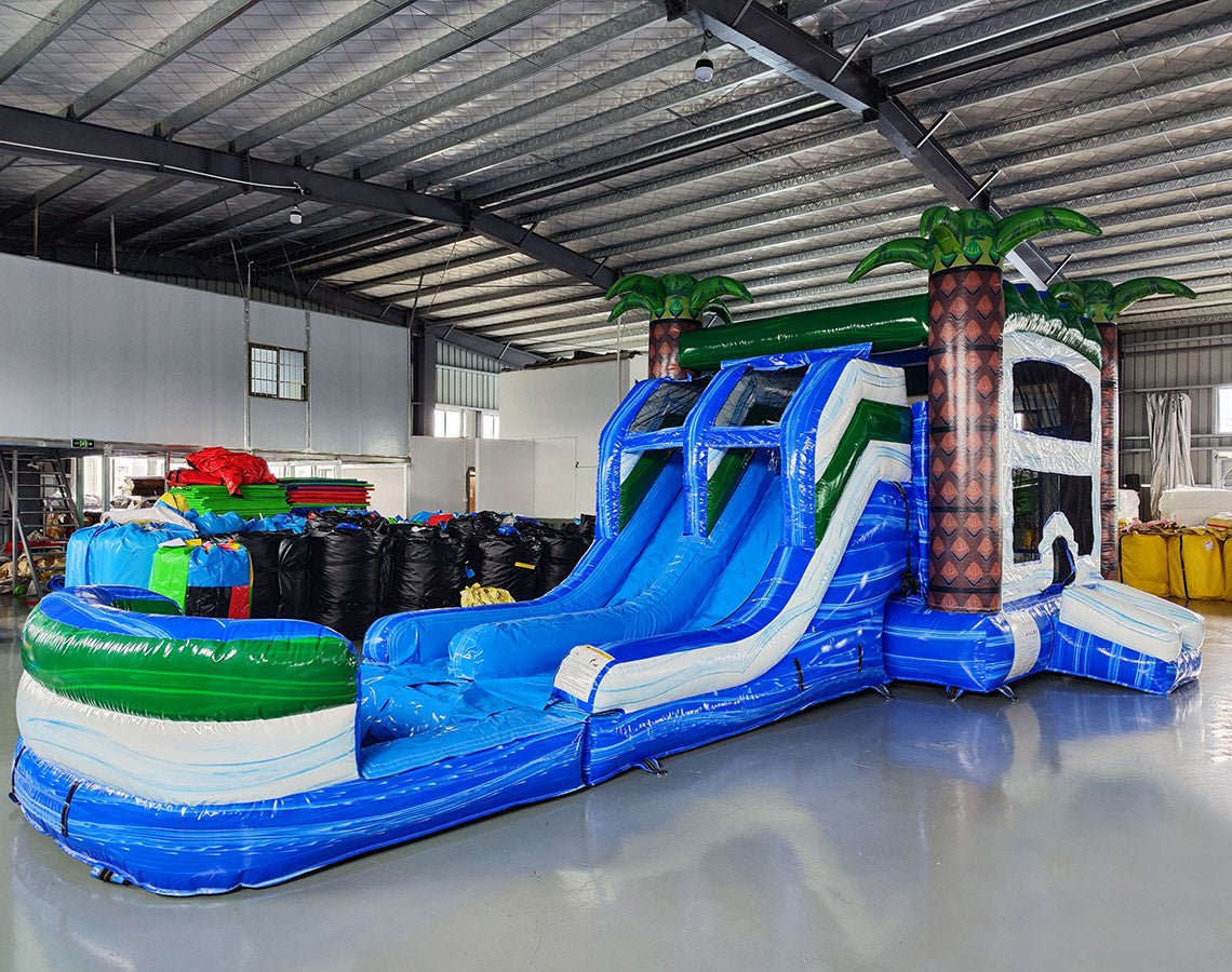 Side view of Island Thunder Dual Lane Commercial Inflatable Combo (Wet/Dry) showing dual blue water slides, inflatable palm trees with brown trunks and green leaves, and a large blue splash pool. The combo includes a white bounce house area and green accents. Set in a warehouse with metal ceiling and various inflatable products visible.