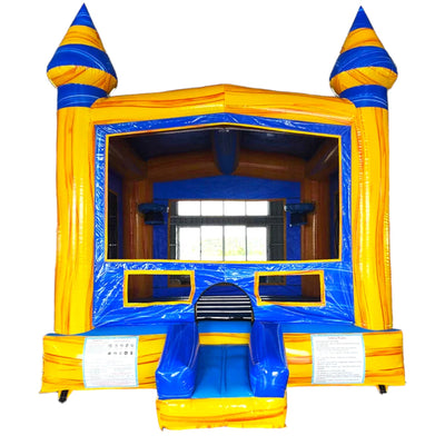 Large inflatable Lava Falls Commercial Grade Bounce House featuring a vibrant volcano theme. The structure has a blue base, green pillars resembling palm trees, and a yellow and blue top section with cone-shaped turrets. A curved yellow slide emerges from one side. The bounce area has mesh windows for visibility and safety. The inflatable is set up in a spacious indoor warehouse with metal roofing and fluorescent lighting. Other inflatable products and equipment are visible in the background.