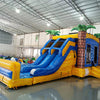 Three-quarter view of Lava Falls Dual Lane Commercial Inflatable Combo set up in an indoor facility. The structure displays a volcano theme with dual blue water slides, orange and yellow lava-like designs, and inflatable palm trees. A blue bounce house area and splash pool are prominent. The warehouse setting shows metal roofing, support beams, and other inflatable products, showcasing its commercial application.