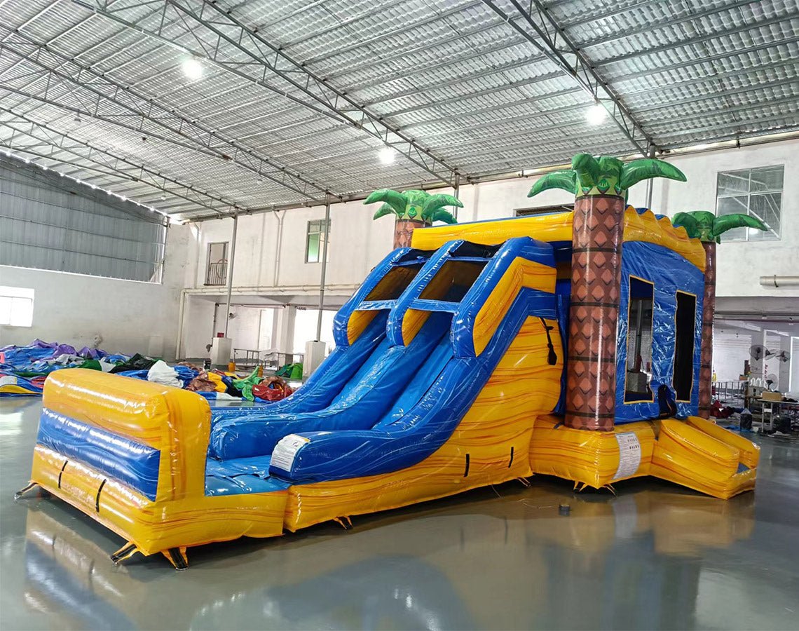 Three-quarter view of Lava Falls Dual Lane Commercial Inflatable Combo set up in an indoor facility. The structure displays a volcano theme with dual blue water slides, orange and yellow lava-like designs, and inflatable palm trees. A blue bounce house area and splash pool are prominent. The warehouse setting shows metal roofing, support beams, and other inflatable products, showcasing its commercial application.