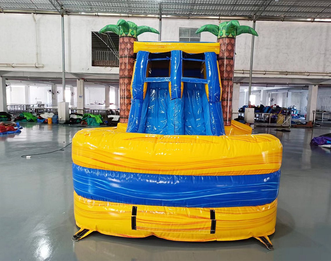 Front view of Lava Falls Dual Lane Commercial Inflatable Combo showing dual blue water slides emerging from an orange and yellow volcano-themed structure. Inflatable palm trees flank the sides, and a blue bounce house area is visible. The inflatable is set against a white background, highlighting its tropical volcano design for commercial wet/dry use.