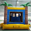 Rear view of Lava Falls Dual Lane Commercial Inflatable Combo (Wet/Dry) featuring a vibrant volcano theme. The structure has dual blue water slides emerging from an orange and yellow lava-patterned base. Inflatable palm trees with green leaves flank the sides. A blue bounce house area with mesh windows is visible in the center, surrounded by the orange and yellow volcano design. Set up in a large indoor warehouse with metal roofing, bright lighting, and other inflatable products visible in the background.