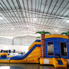 Wide-angle view of Lava Falls Dual Lane Commercial Inflatable Combo in a spacious warehouse. The inflatable features a volcano design with dual blue slides, orange and yellow lava patterns, and palm trees. A blue bounce house and splash pool area are visible. Various other inflatable products and warehouse equipment can be seen, demonstrating its size and commercial setting.