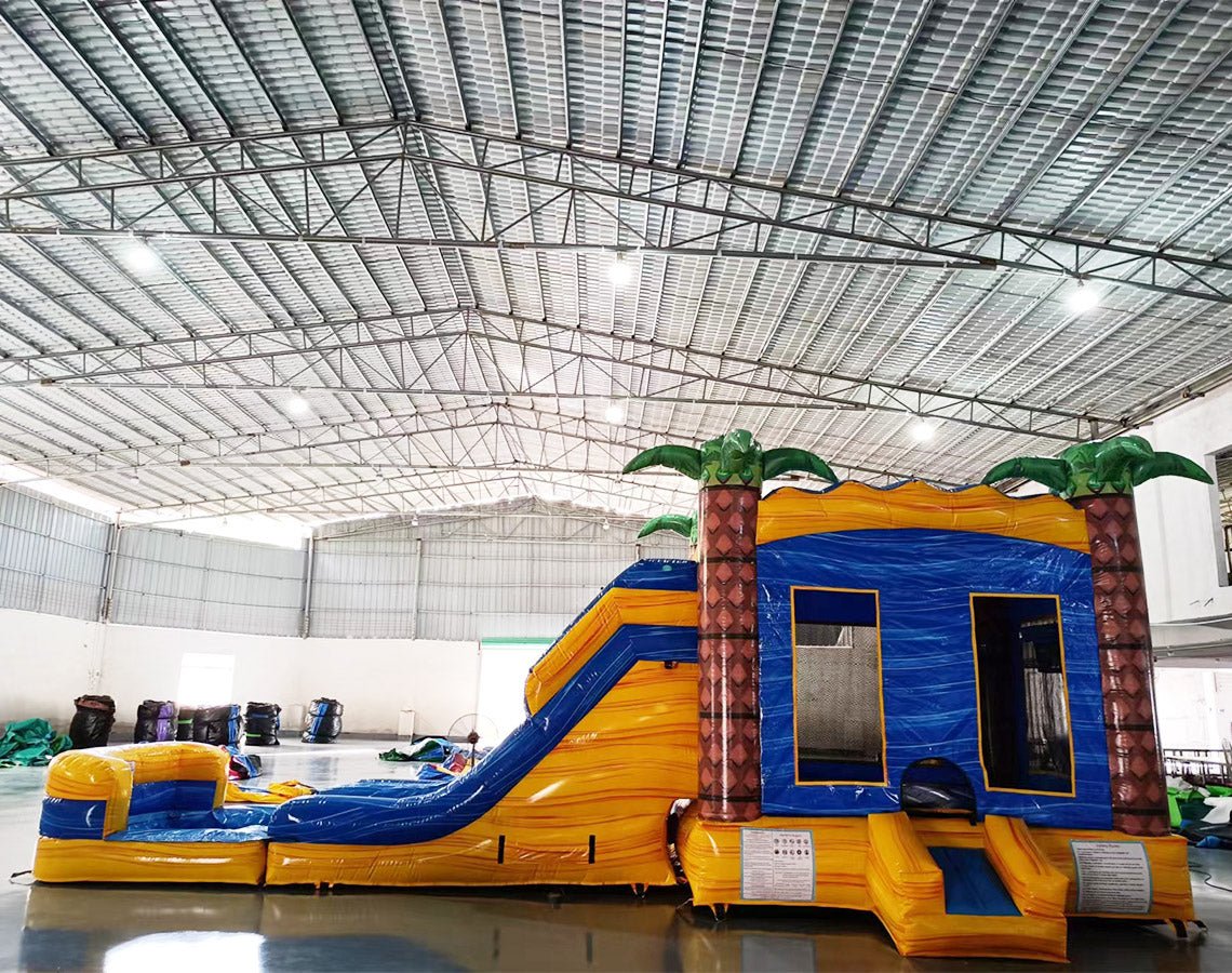 Wide-angle view of Lava Falls Dual Lane Commercial Inflatable Combo in a spacious warehouse. The inflatable features a volcano design with dual blue slides, orange and yellow lava patterns, and palm trees. A blue bounce house and splash pool area are visible. Various other inflatable products and warehouse equipment can be seen, demonstrating its size and commercial setting.