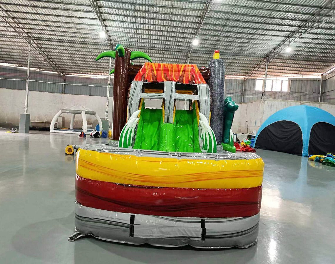 Front view of Lava Raptor Dual Lane Commercial Inflatable Combo featuring a prehistoric theme. The structure has dual slides, one red and one green, emerging from a brown volcano-like base with orange lava patterns. Two large inflatable dinosaurs flank the entrance. Set up in a spacious warehouse with metal roofing and bright lighting visible.