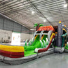 Wide-angle view of Lava Raptor Dual Lane Commercial Inflatable Combo in a large indoor facility. The inflatable displays a volcano design with dual slides, dinosaur figures, and a splash pool area. Various inflatable obstacles and decorations are visible. The warehouse setting shows metal roofing, support beams, and other equipment, demonstrating its size and commercial application.