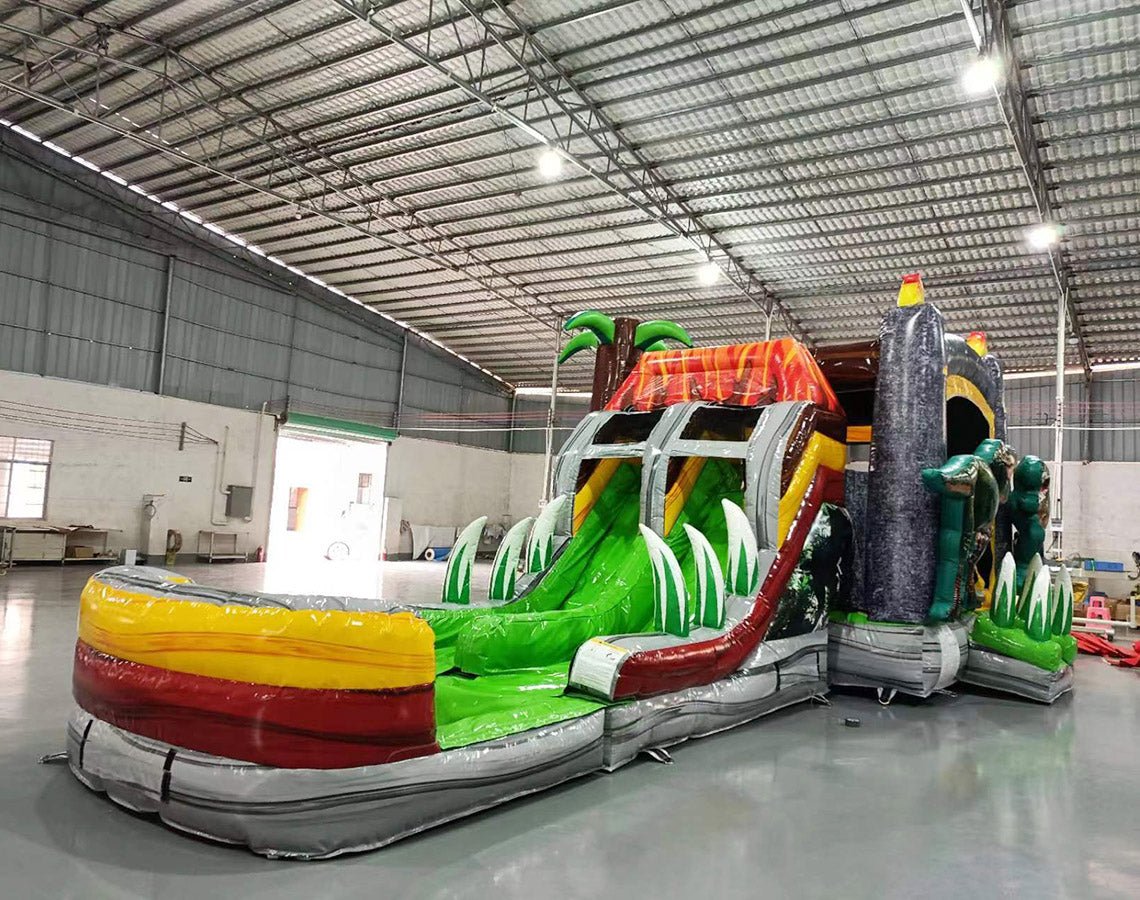 Wide-angle view of Lava Raptor Dual Lane Commercial Inflatable Combo in a large indoor facility. The inflatable displays a volcano design with dual slides, dinosaur figures, and a splash pool area. Various inflatable obstacles and decorations are visible. The warehouse setting shows metal roofing, support beams, and other equipment, demonstrating its size and commercial application.