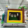 Interior view of Lava Raptor Dual Lane Commercial Inflatable Combo (Wet/Dry) showing the entrance and bounce area. The inflatable features a yellow archway entrance with brown tree trunk designs on either side. Green inflatable palm leaves are visible at the top. The interior shows a green bounce floor and inflatable obstacles. Set in a warehouse with concrete flooring and various other inflatable products visible in the background.