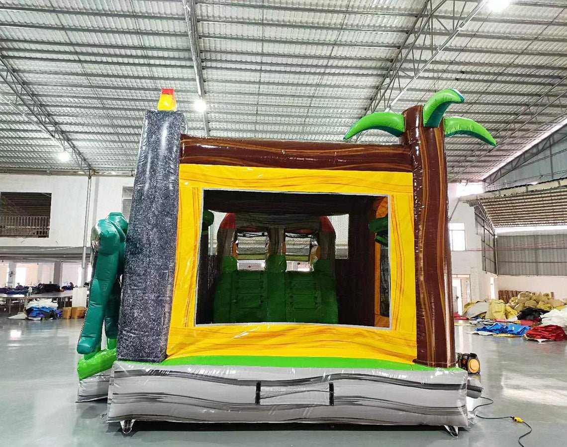 Interior view of Lava Raptor Dual Lane Commercial Inflatable Combo (Wet/Dry) showing the entrance and bounce area. The inflatable features a yellow archway entrance with brown tree trunk designs on either side. Green inflatable palm leaves are visible at the top. The interior shows a green bounce floor and inflatable obstacles. Set in a warehouse with concrete flooring and various other inflatable products visible in the background.