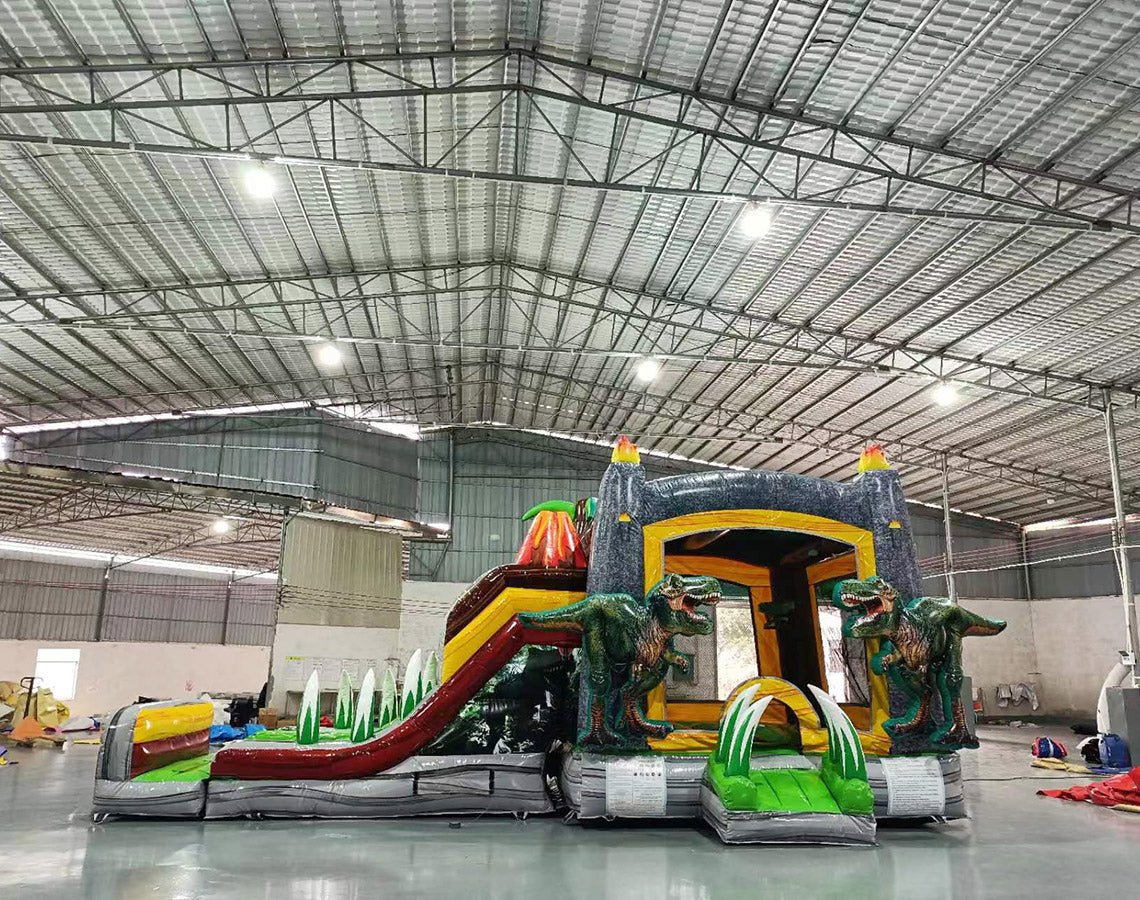 Three-quarter view of Lava Raptor Dual Lane Commercial Inflatable Combo set up in a warehouse. The structure features a brown volcano base with orange lava patterns, dual slides, and large dinosaur figures at the entrance. A colorful splash pool and various inflatable obstacles are visible. The image showcases the combo's size and detailed prehistoric theme in a commercial setting.