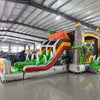 Front view of Lava Raptor Dual Lane Commercial Inflatable Combo (Wet/Dry) featuring a prehistoric volcano theme. The structure has dual slides, one red and one green, emerging from a brown volcano-like base with orange lava patterns. Two large inflatable dinosaur figures flank the entrance. A colorful splash pool area is visible at the base. Set up in a spacious warehouse with metal roofing and bright lighting visible.