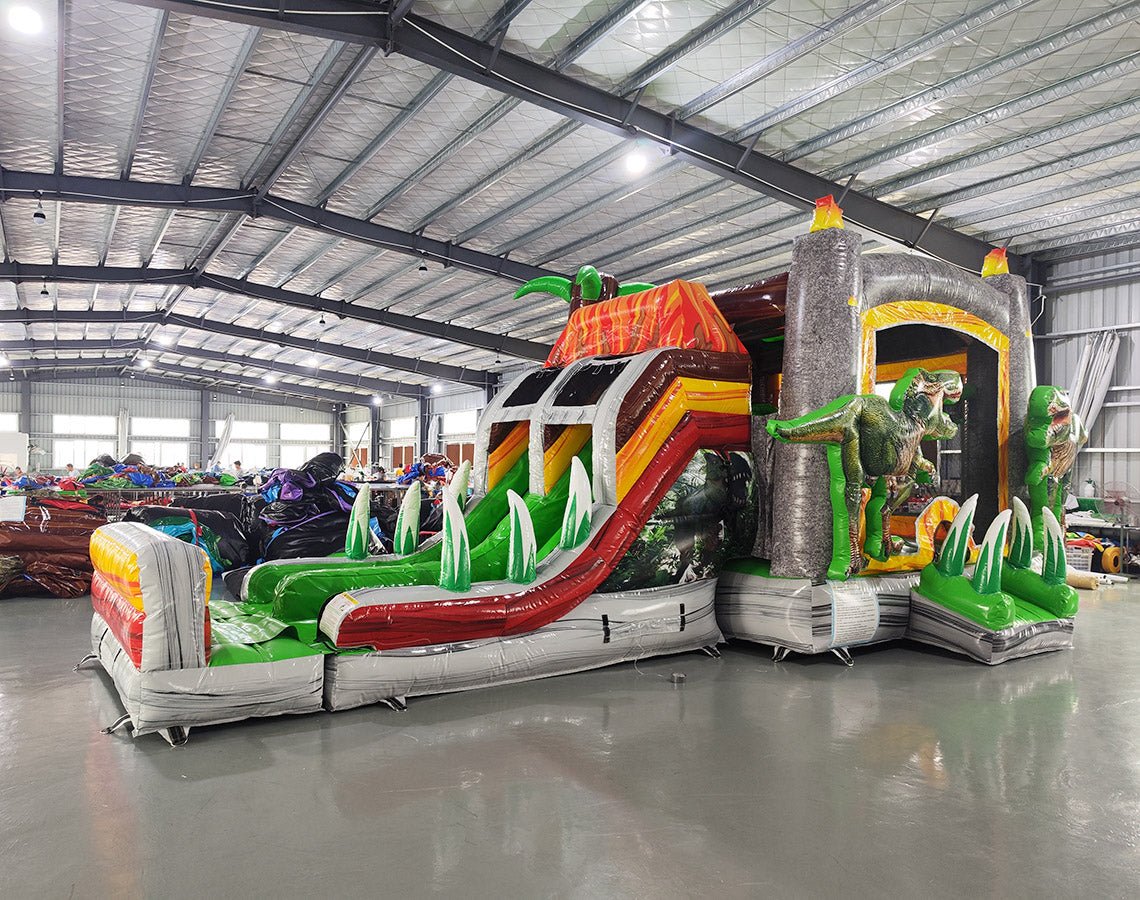 Front view of Lava Raptor Dual Lane Commercial Inflatable Combo (Wet/Dry) featuring a prehistoric volcano theme. The structure has dual slides, one red and one green, emerging from a brown volcano-like base with orange lava patterns. Two large inflatable dinosaur figures flank the entrance. A colorful splash pool area is visible at the base. Set up in a spacious warehouse with metal roofing and bright lighting visible.