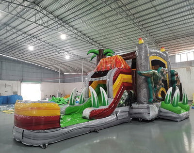 Wide-angle view of the Lava Raptor Econo Combo featuring a volcano-shaped bounce house with lava design, red slide, splash pool, and multiple realistic dinosaur figures. Set up in a spacious indoor warehouse with metal roofing, bright lighting, and other inflatable products visible in the background.