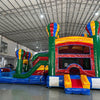 Large inflatable combo featuring a colorful bounce house with hot air balloon designs, attached slide, and splash pool. The structure has vibrant red, blue, green, and yellow colors with multiple balloon-shaped towers. Set up in an indoor warehouse with metal roofing visible.