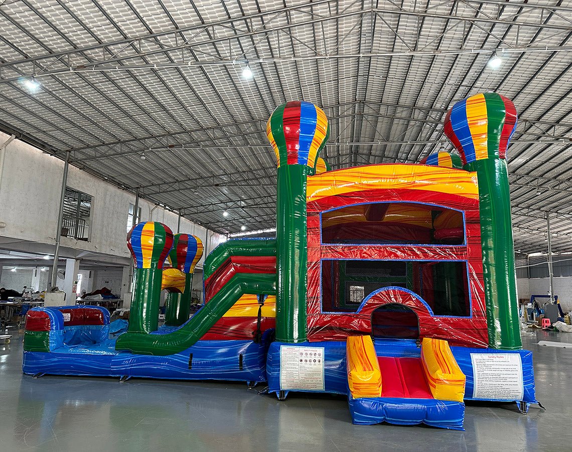 Large inflatable combo featuring a colorful bounce house with hot air balloon designs, attached slide, and splash pool. The structure has vibrant red, blue, green, and yellow colors with multiple balloon-shaped towers. Set up in an indoor warehouse with metal roofing visible.