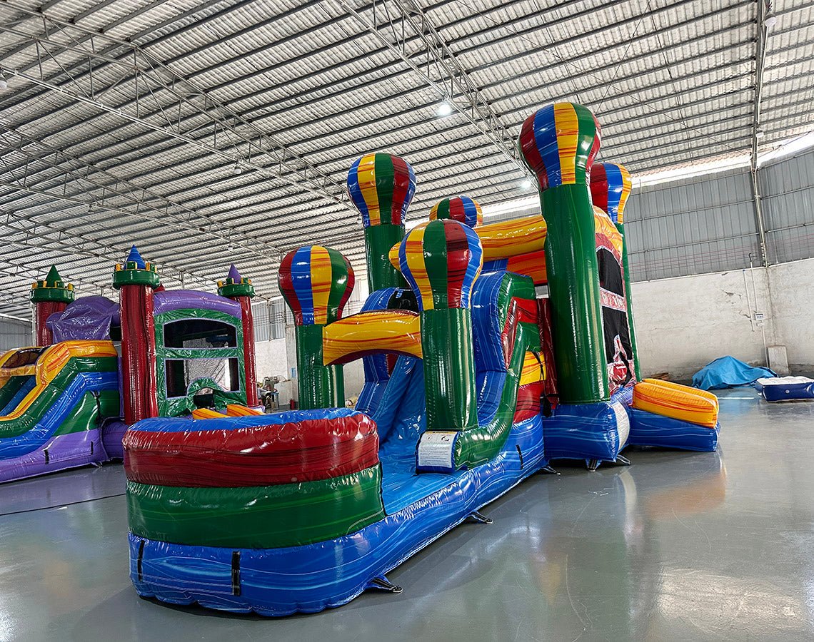 Wide-angle view of Marble Balloon Econo Combo featuring multiple hot air balloon-shaped towers, bounce house, slides, and splash pool area. The inflatable is set up in a spacious indoor warehouse with metal roofing, bright lighting, and other inflatable products visible in the background.