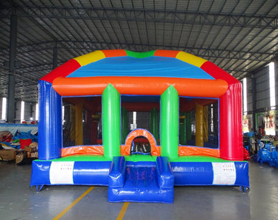 Front angle view of Mega Fun Dome Commercial Grade Bounce House in a warehouse setting. The inflatable structure has a blue domed roof with multicolored panels, orange support beams, and green entrance pillars. A small orange archway marks the entrance. The bounce house is surrounded by other inflatable products and warehouse equipment, demonstrating its size and commercial application.