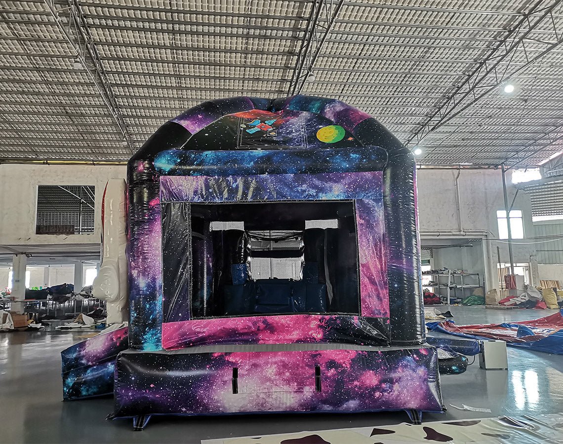 Large inflatable space-themed combo featuring a bounce house with cosmic design, attached slide, and play area. The structure has vibrant purple, blue, and pink colors with astronaut and rocket decorations. Set up in an indoor warehouse with metal roofing visible.