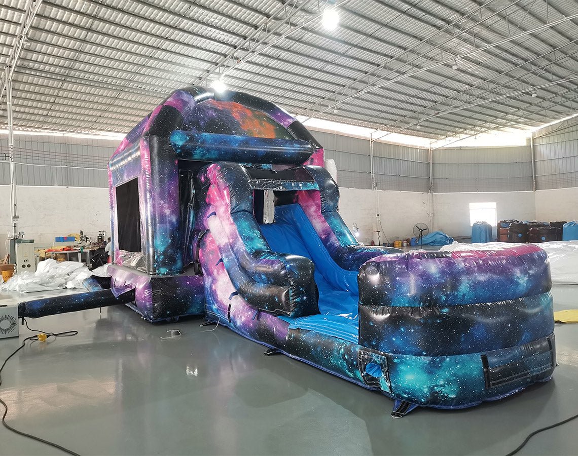 Side angle view of MoonWalker Commercial Econo Inflatable Wet/Dry Combo showing space-themed inflatable with cosmic patterns, slide, and play area. Features vibrant purple, blue, and pink colors with astronaut and rocket designs. Set up in a spacious indoor warehouse with metal roofing visible.