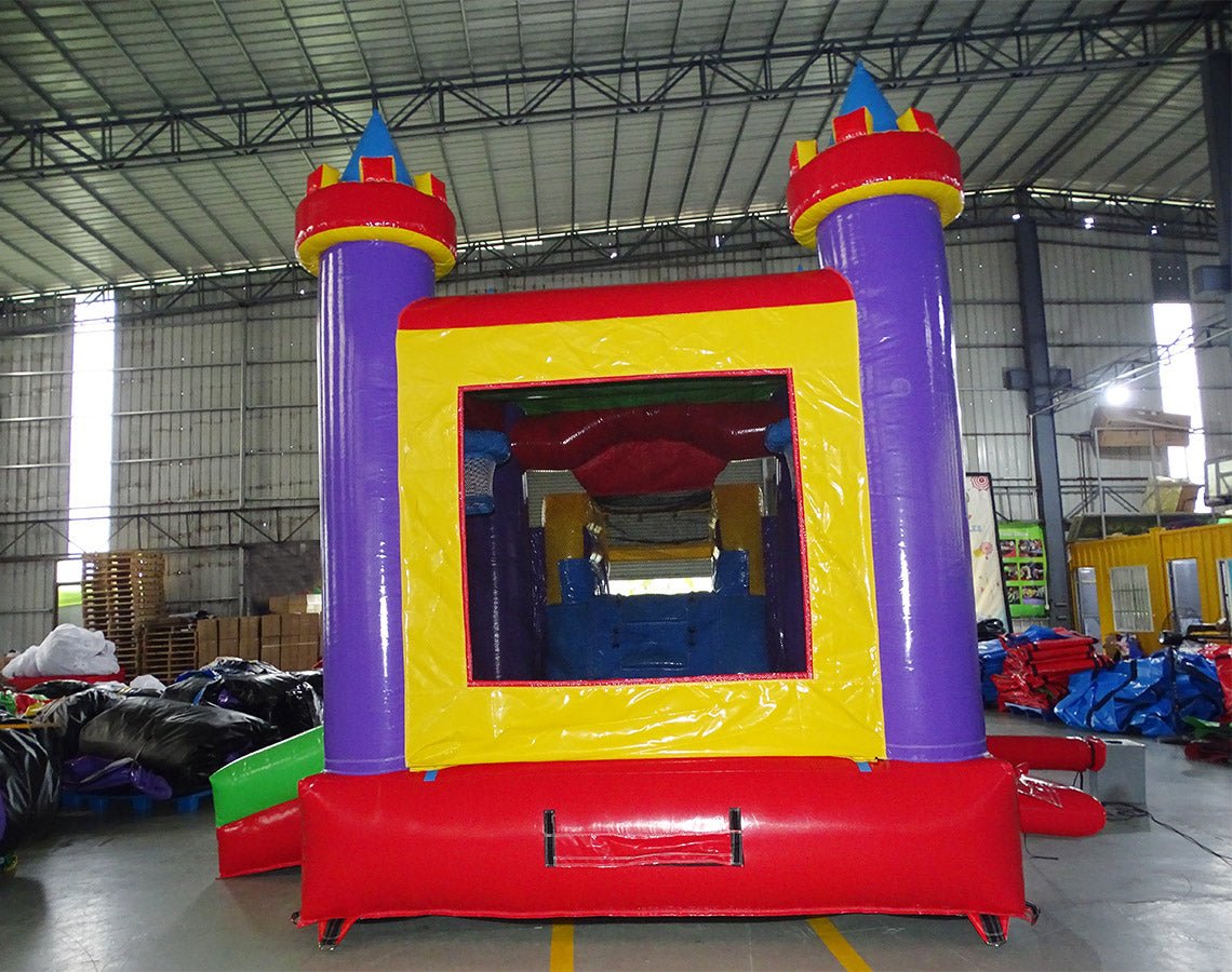 Rear view of Multi Color Castle Commercial Econo Inflatable Combo (Wet/Dry) showing the bounce house entrance with purple turrets and yellow frame. Interior visible through mesh windows. Set up in an indoor warehouse with other inflatable products and equipment in the background.