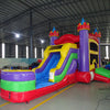 Side view of Multi Color Castle Commercial Econo Inflatable Combo (Wet/Dry) set up in a large indoor warehouse. Features purple turrets, yellow walls, blue slide, green obstacles, and red splash pool. Other inflatable products and warehouse equipment visible in the background.