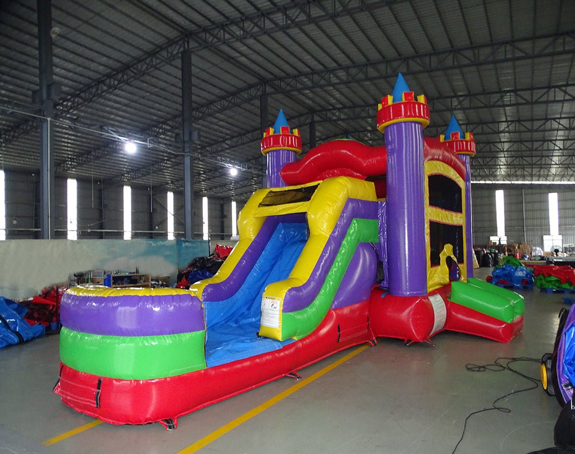 Side view of Multi Color Castle Commercial Econo Inflatable Combo (Wet/Dry) set up in a large indoor warehouse. Features purple turrets, yellow walls, blue slide, green obstacles, and red splash pool. Other inflatable products and warehouse equipment visible in the background.