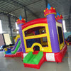 Three-quarter view of Multi Color Castle Commercial Econo Inflatable Combo (Wet/Dry) showcasing purple turrets, yellow walls, blue slide, green obstacles, and red base. Inflatable displayed in a large indoor warehouse with metal roofing and other equipment visible.