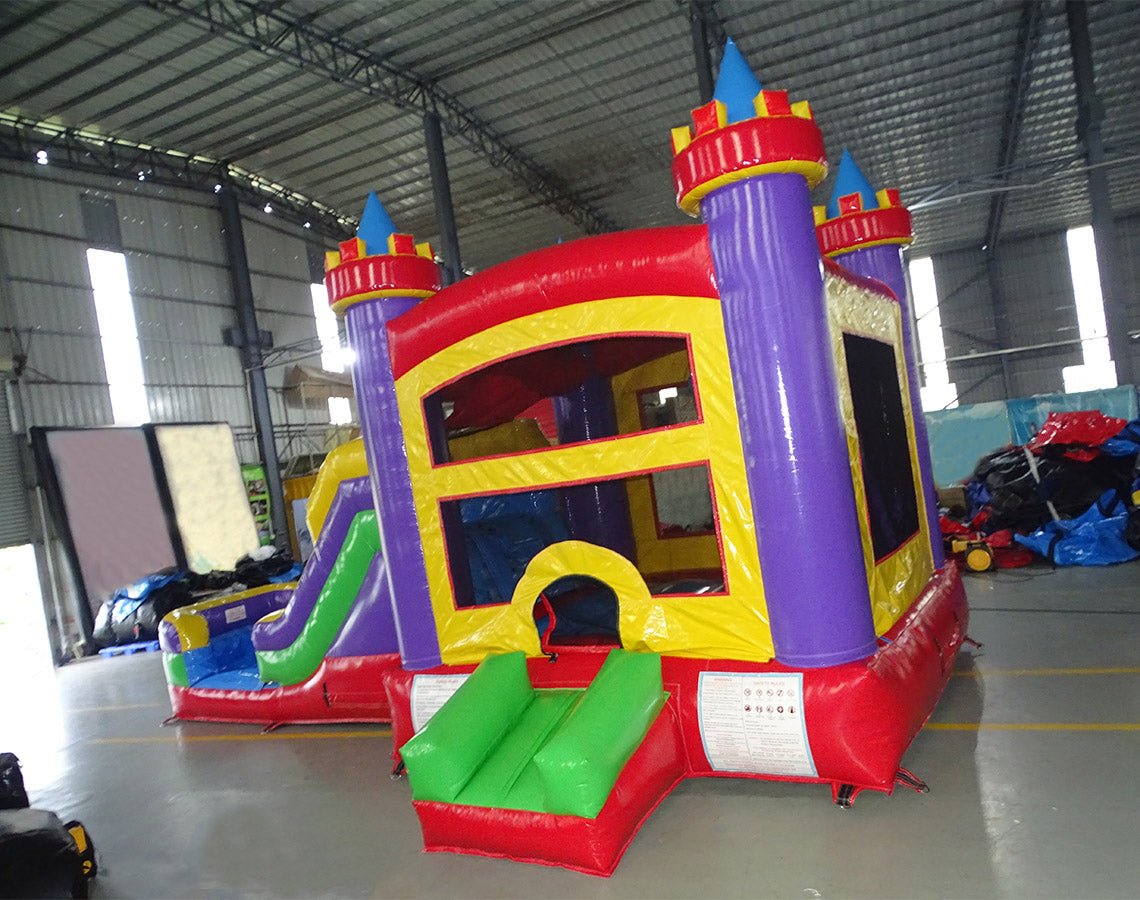 Three-quarter view of Multi Color Castle Commercial Econo Inflatable Combo (Wet/Dry) showcasing purple turrets, yellow walls, blue slide, green obstacles, and red base. Inflatable displayed in a large indoor warehouse with metal roofing and other equipment visible.