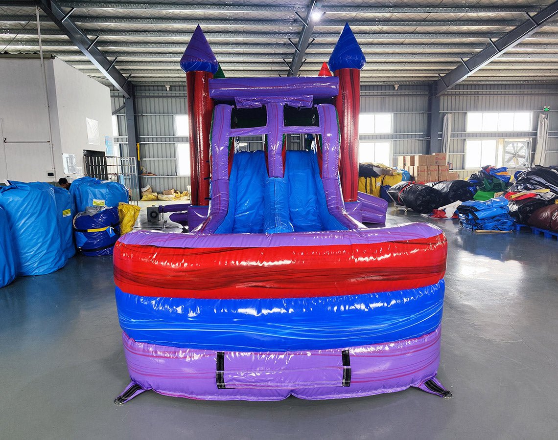 Front view of Multi Color Marble Splash & Save Dual Lane Inflatable Combo showing dual blue slides, purple castle towers with conical tops, and a colorful splash pool area. Set up in a warehouse with concrete flooring, metal roofing, and various other inflatable products and equipment visible in the background.