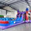 Wide-angle view of Multi Color Marble Splash & Save Dual Lane Inflatable Combo in a spacious warehouse. The inflatable displays vibrant purple, blue, and red colors with castle towers, dual slides, and a large splash pool. Various inflatable products, storage bags, and warehouse equipment are visible, demonstrating its size and commercial setting.